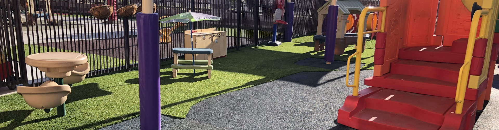 Synthetic grass installation at a children's playground with rubber safety surfacing, colorful climbing structures, activity tables, and a black iron perimeter fence